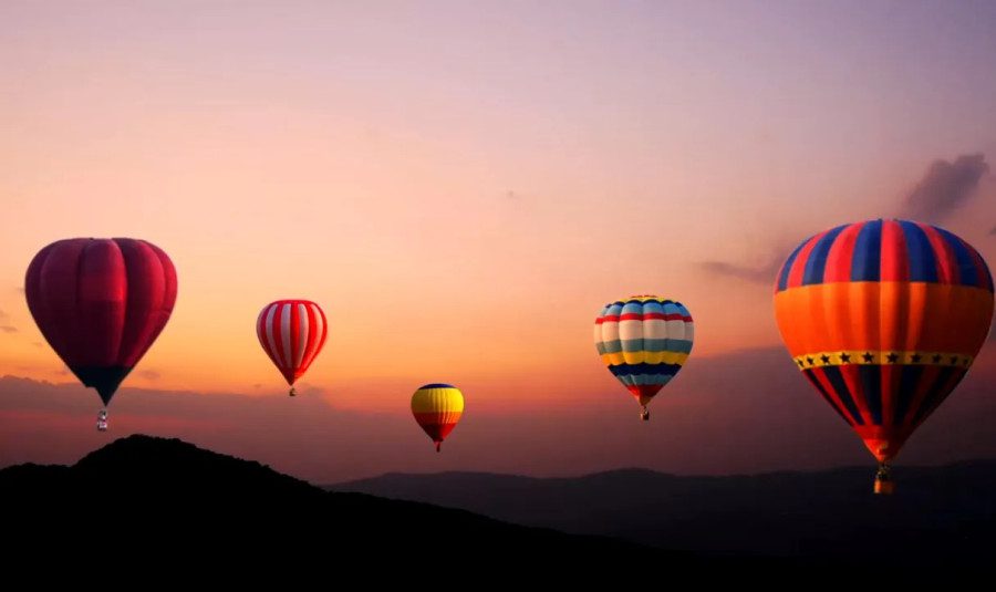 Five hot air balloons of various colors and patterns float across a gradient sunset sky above silhouetted mountains.