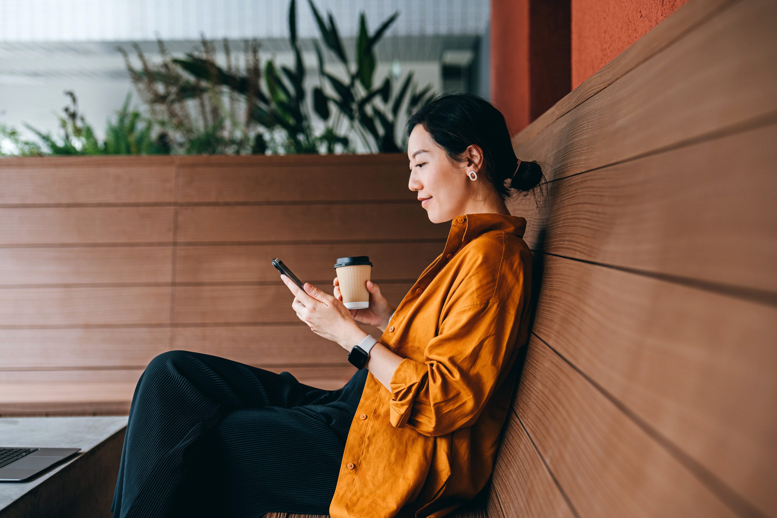 Young Asian woman sitting in a sidewalk cafe with a cup of coffee and using her smartphone. Enjoying a relaxing moment in the afternoon. Coffee break. Lifestyle and technology