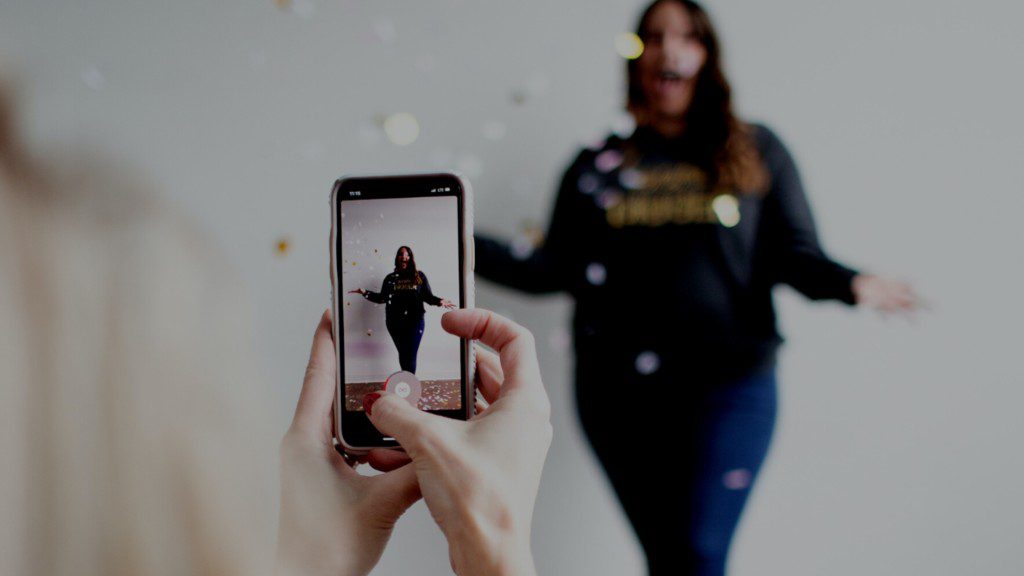 In the scene, a woman is capturing a photo of another woman with confetti in the background.