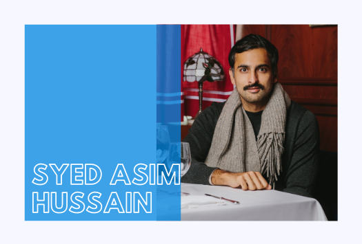 Syed Asim Hussain sitting at a table and wearing a grey scarf and dark grey top