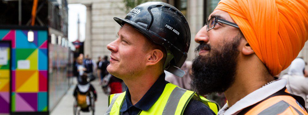 Two construction workers. One wears a hard hat and one wears a turban, both wear high-vis jackets with an Erith logo emblazoned.