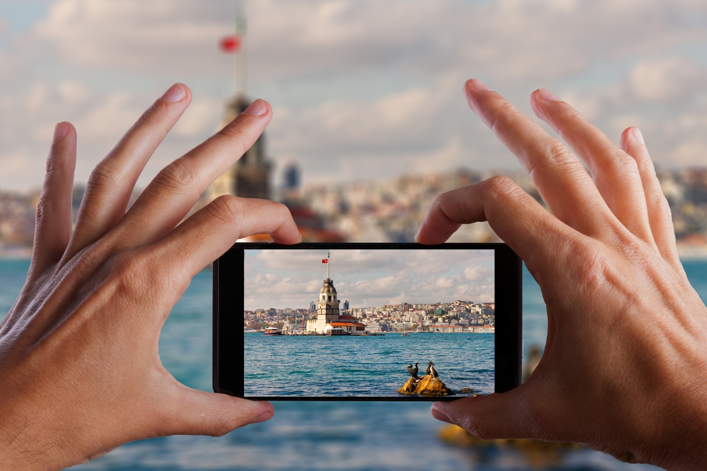 A smartphone held by two hands captures a photo of the Maiden's Tower in Istanbul, Turkey, with the Bosphorus Strait in the foreground.