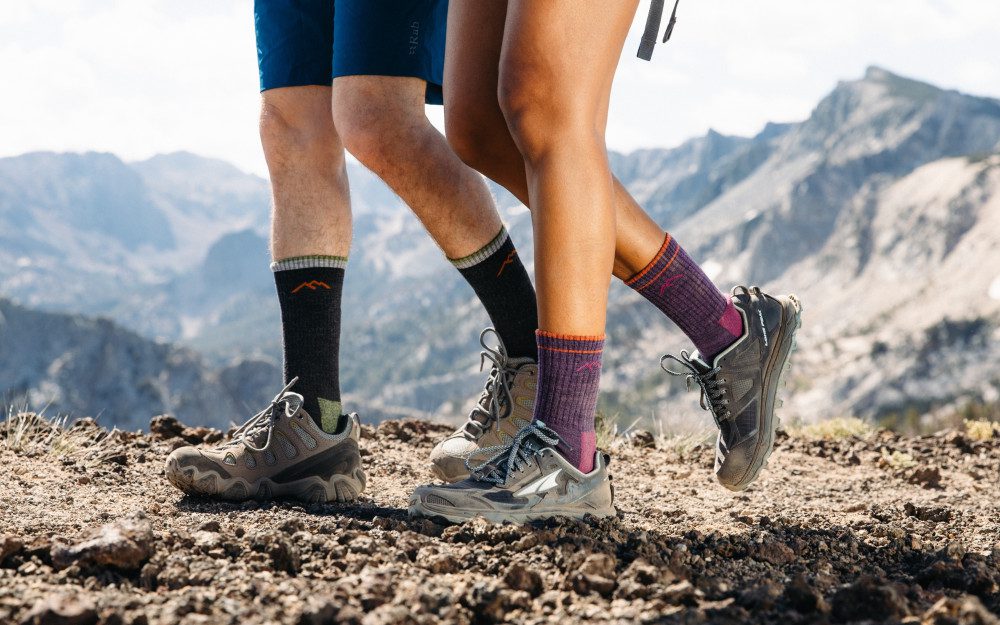 Hikers in hiking socks and trail shoes step over rocky ground along a mountain ridge, with blue-gray peaks and a cloudy sky in the background.