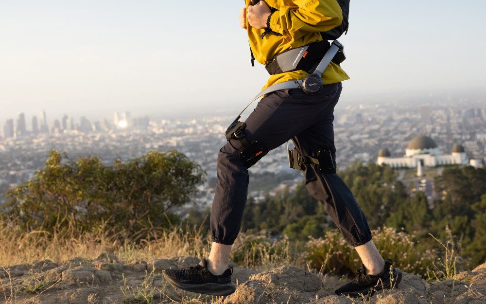 A hiker in a yellow jacket and dark pants walks across rocky ground on a hilltop, wearing a backpack and looking over a sprawling city skyline in the distance.
